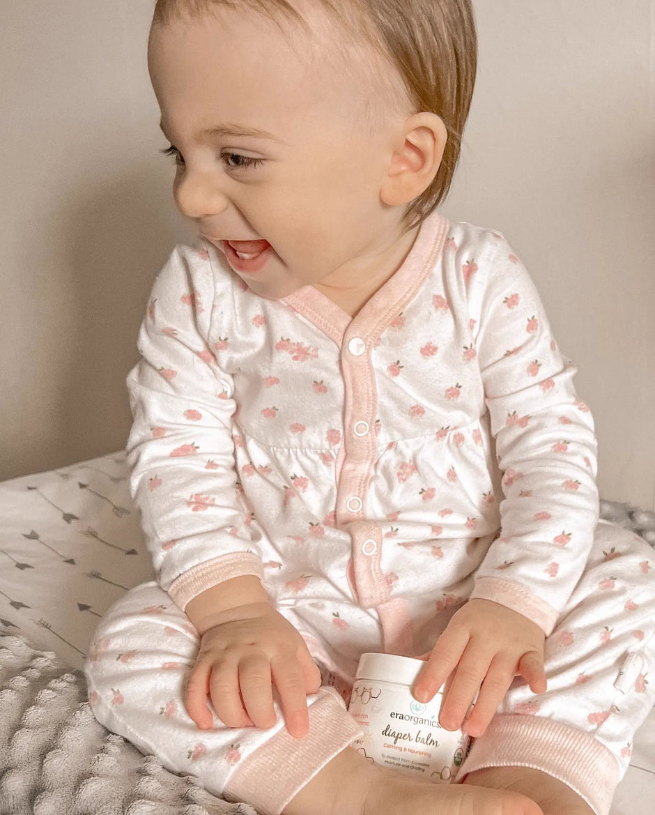 Baby in pink pajamas holding a jar of Diaper Balm
